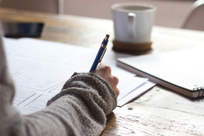 Person writing on paper on a desk with mug in the background